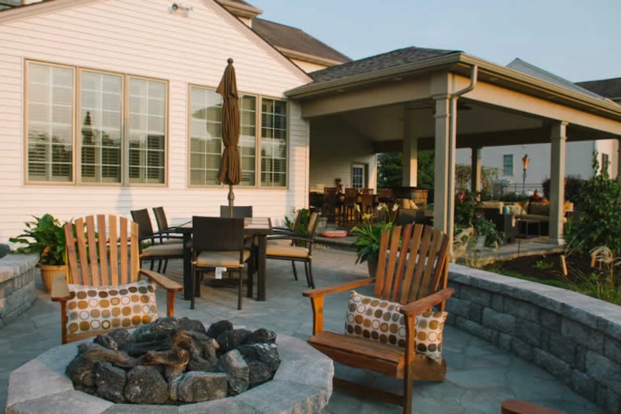 Outdoor patio with Adirondack chairs, fire pit, umbrella, and dining set, adjacent to a house with covered porch.