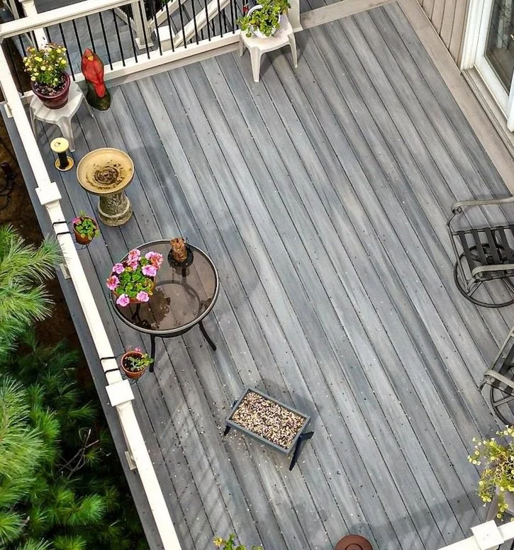 Outdoor deck with gray wooden floor, small glass table with pink flowers and ornament, birdbath, lounge chair, bird feeder, and potted plants.