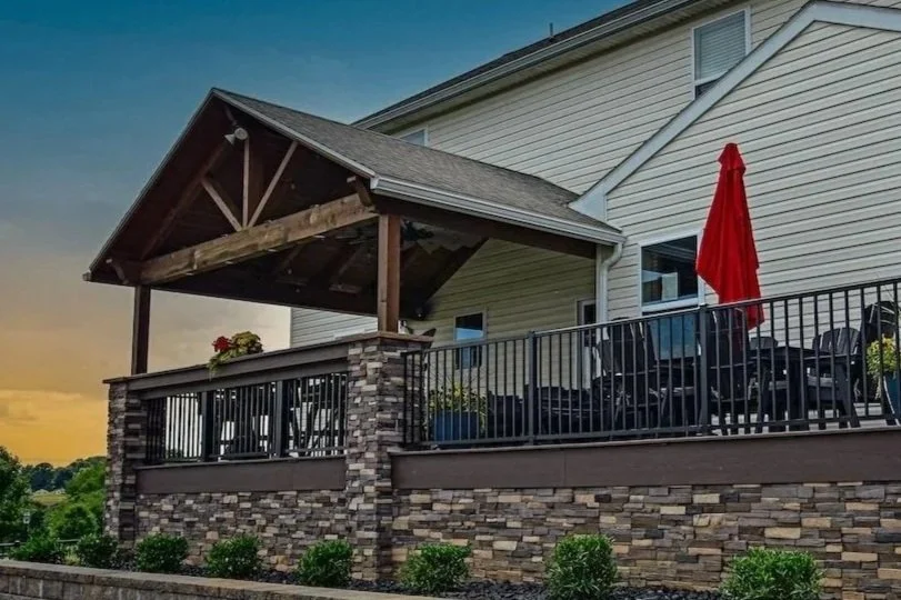 A covered outdoor patio with a gable roof, stone pillars, black metal railing, patio furniture, potted plants, and a red umbrella attached to a house with beige siding.