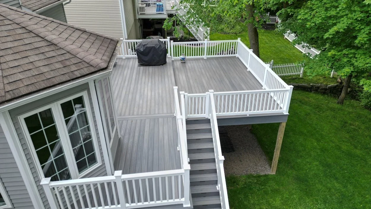 Elevated backyard deck with gray flooring and white railings, featuring stairs leading to a grassy yard and a grill covered with a black cover.