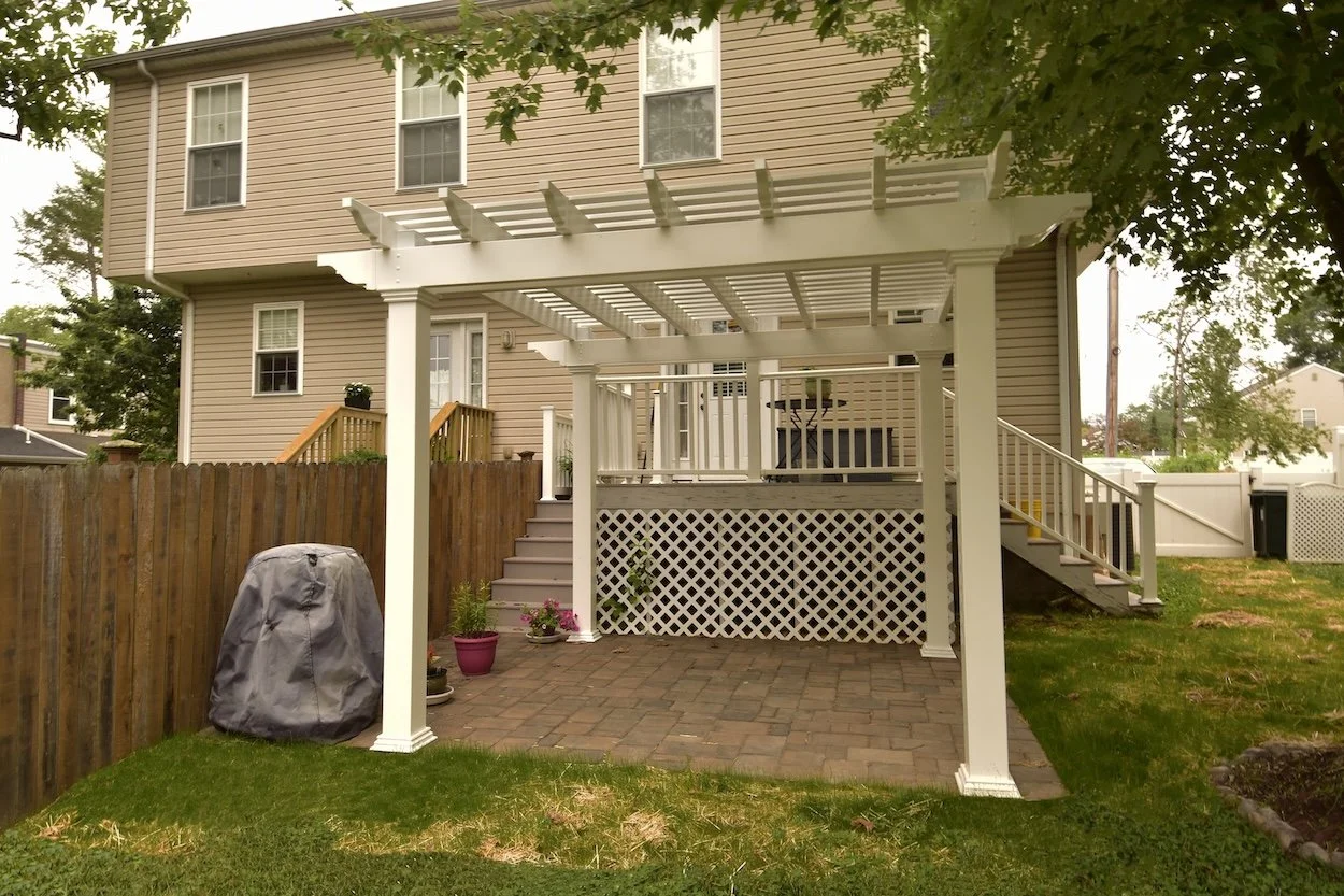 Backyard with white pergola, wooden deck, and patio; house in the background, grass and wooden fence surrounding the area.