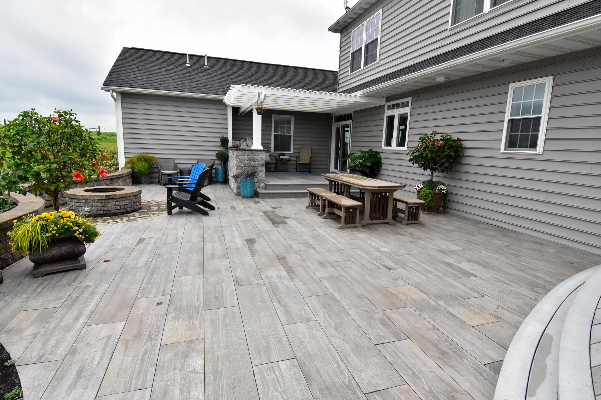 Modern backyard patio with wooden-style tiles, picnic table, chairs, fire pit, plants, and pergola.