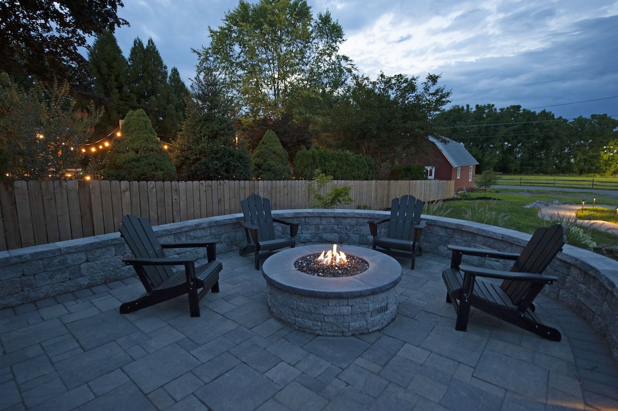 A backyard patio with four black Adirondack chairs arranged around a circular fire pit with small flames, surrounded by a curved stone wall and paving stones, with trees, a wooden fence, a red shed, and string lights in the background during dusk.