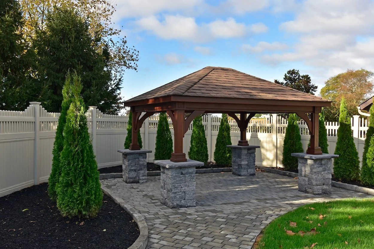 Backyard with a wooden gazebo on stone pillars, surrounded by evergreen trees and a white privacy fence.