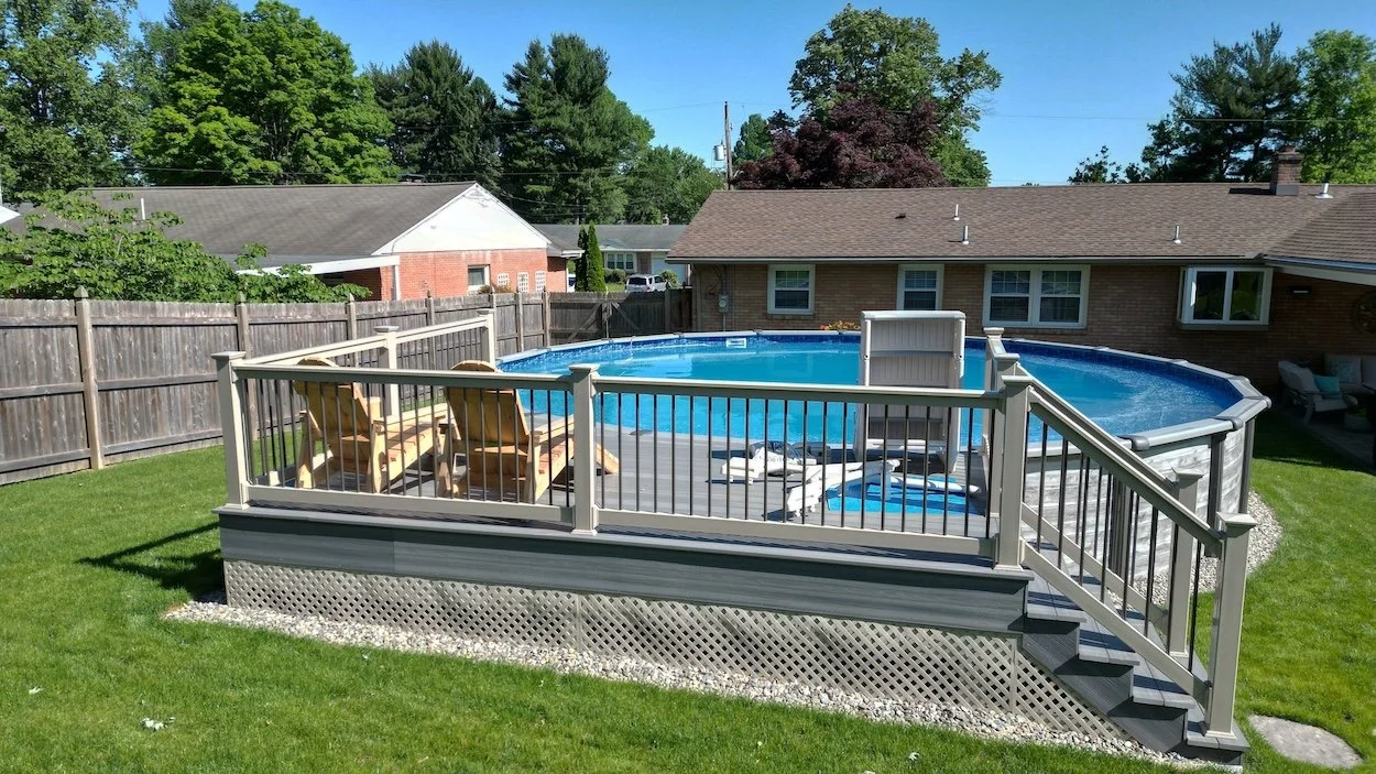 Above-ground pool with wooden deck and chairs in a backyard, surrounded by a wooden fence and lush greenery.