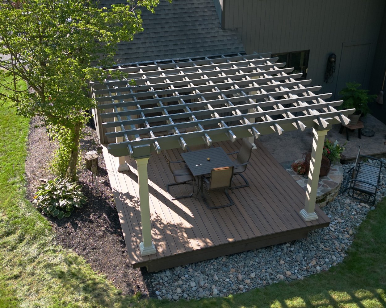 Aerial view of a backyard patio with a wooden pergola and patio furniture. The deck is surrounded by greenery and landscaping, featuring a tree and various plants. The patio area includes a table and chairs for outdoor dining.
