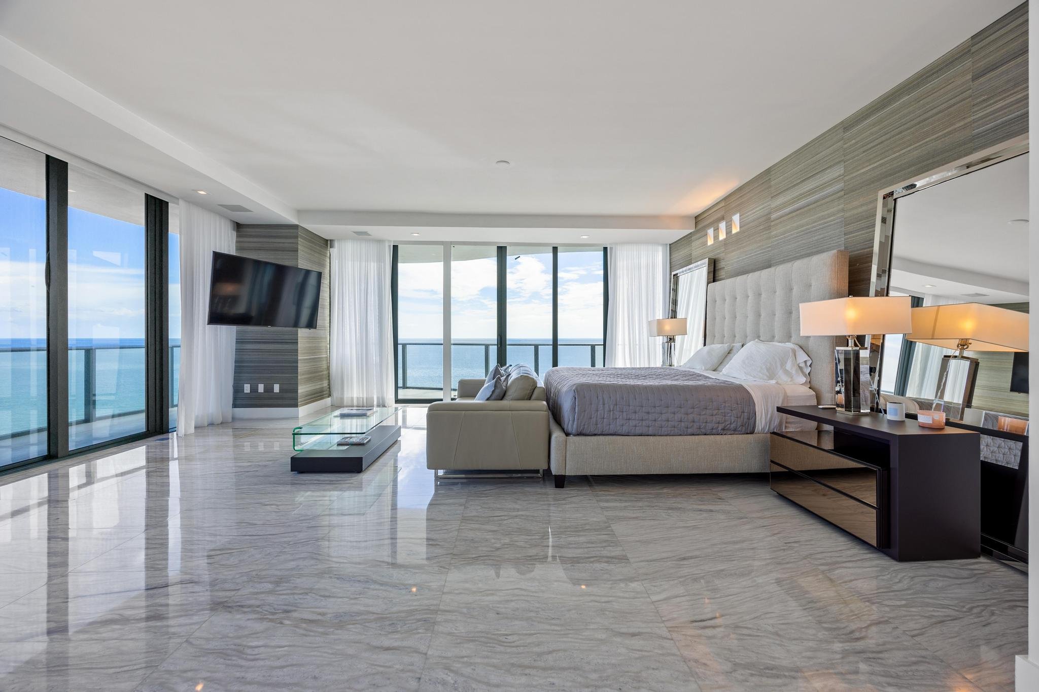 Luxury bedroom with ocean view, featuring a king-size bed, large mirrors, lamps, and modern décor. Floor-to-ceiling windows open to a balcony with glass railing. The room has marble flooring and a wall-mounted flat-screen TV.