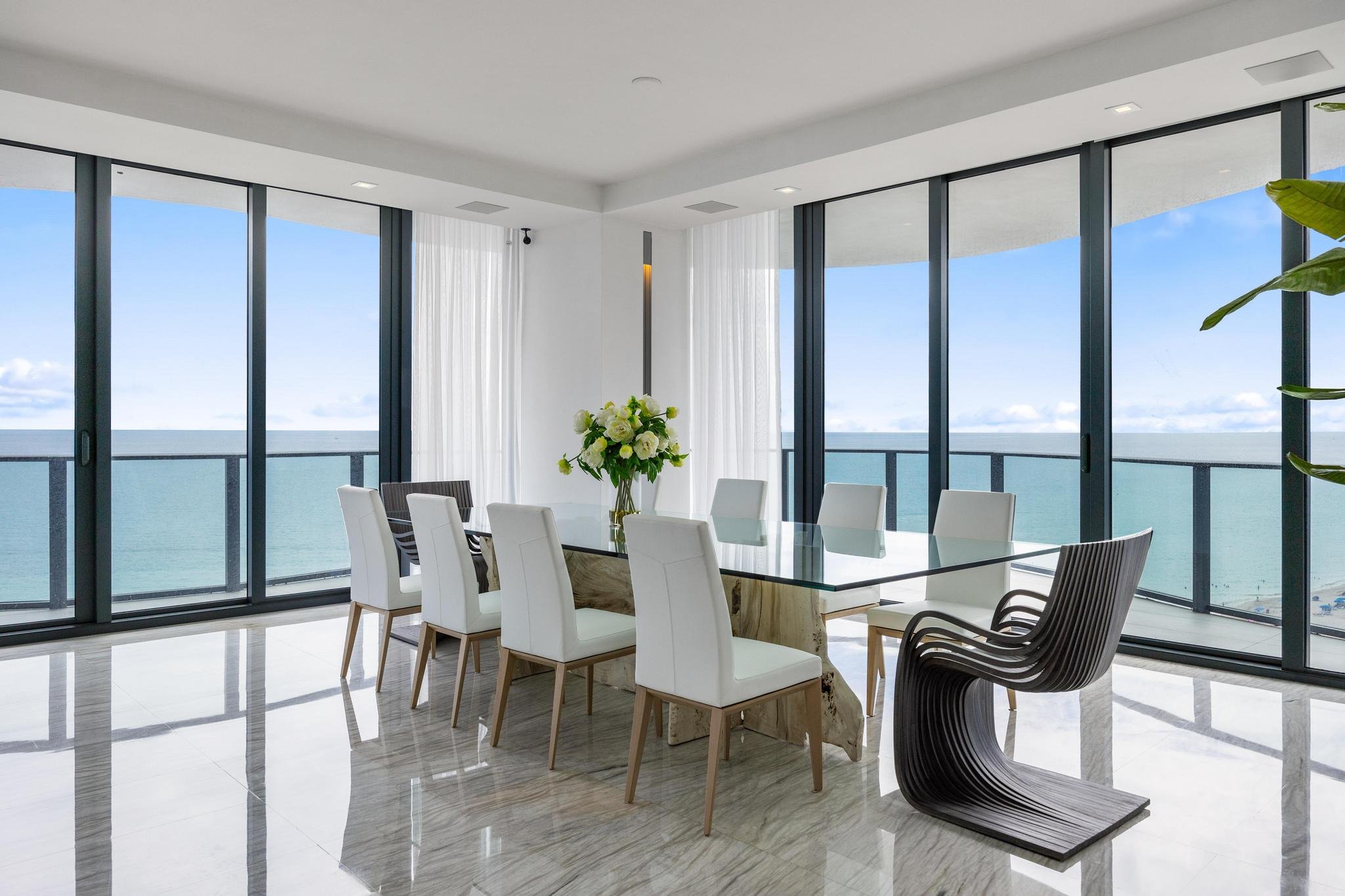 Modern dining room with glass table, white chairs, and ocean view through large windows.