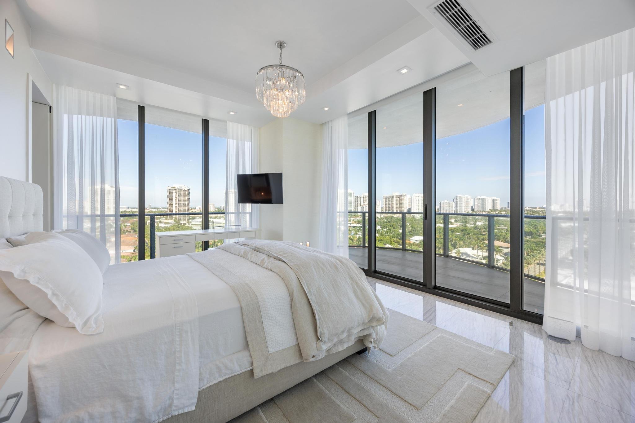 Luxury bedroom with panoramic city views, large windows, white bed linens, modern decor, and a hanging chandelier.