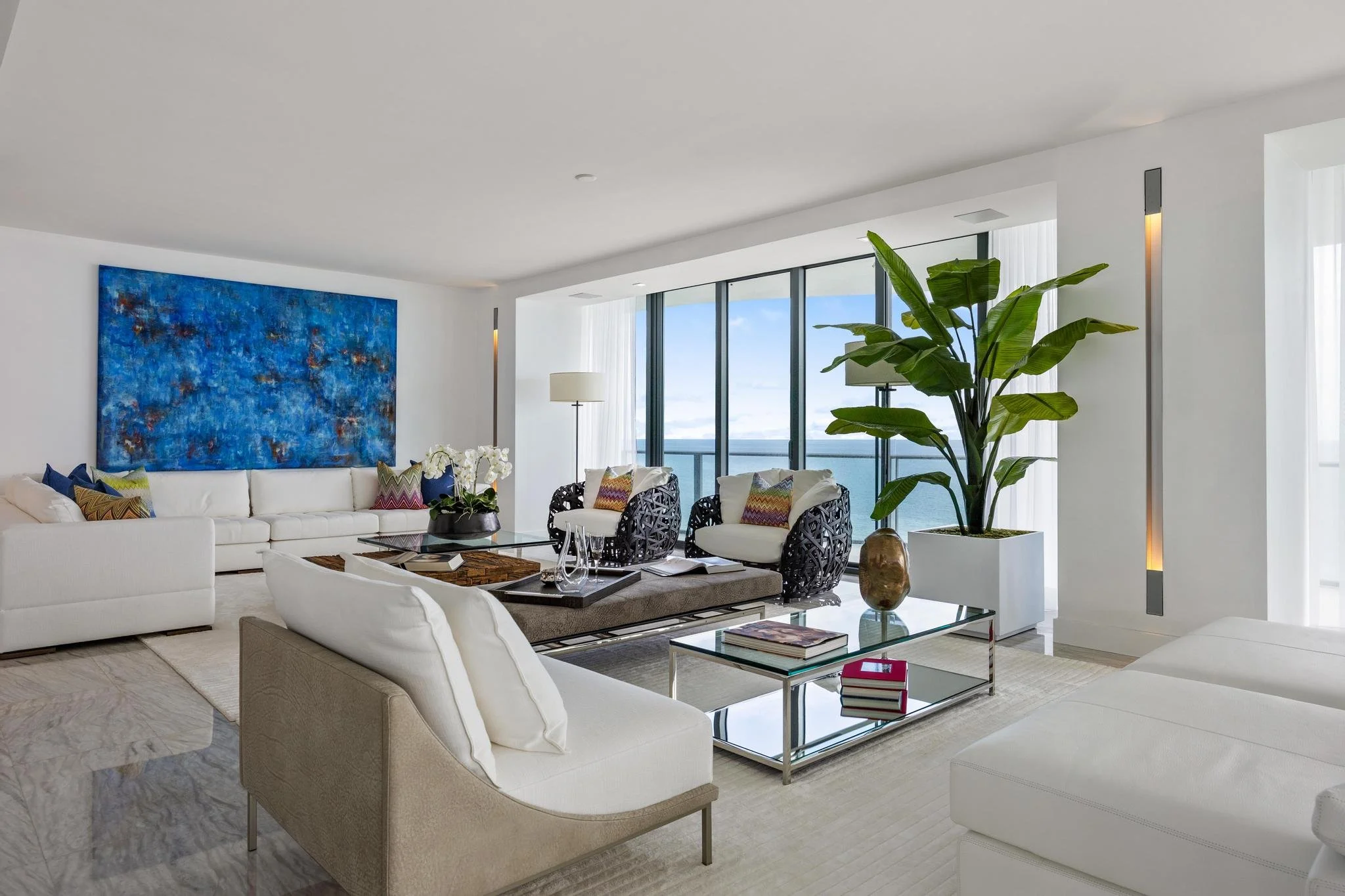 Modern living room with white sofas, large blue abstract painting, black woven armchairs, glass coffee table, floor-to-ceiling windows with ocean view, tall indoor plant, and decorative pillows.