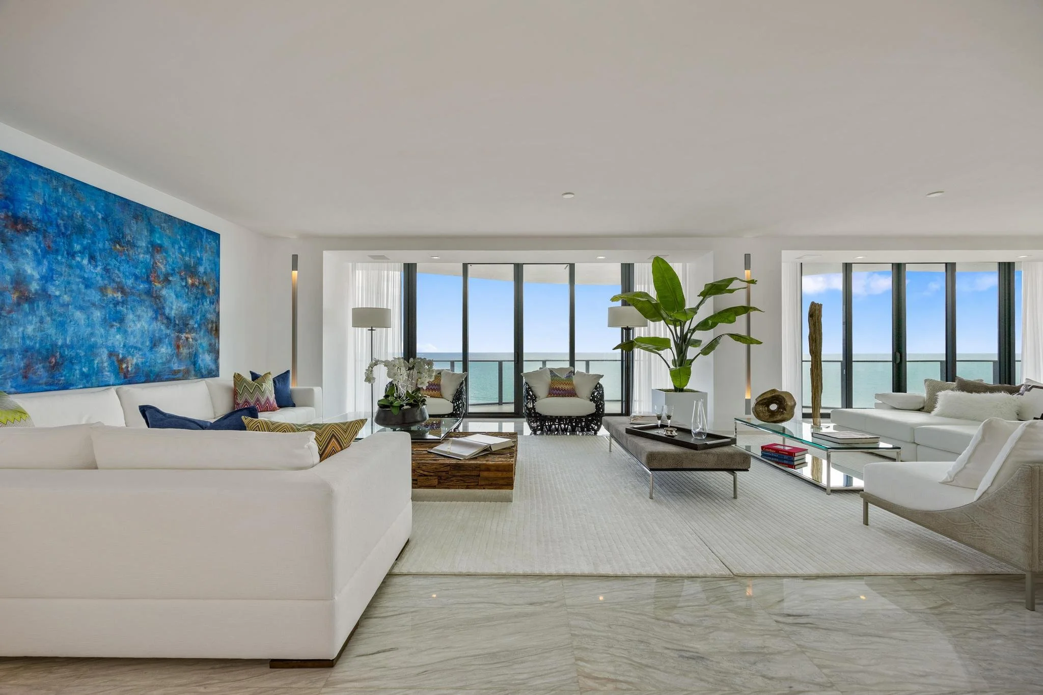 Luxury living room with large windows offering ocean views, featuring white sofas, a blue abstract painting, a glass coffee table, and a large green plant.