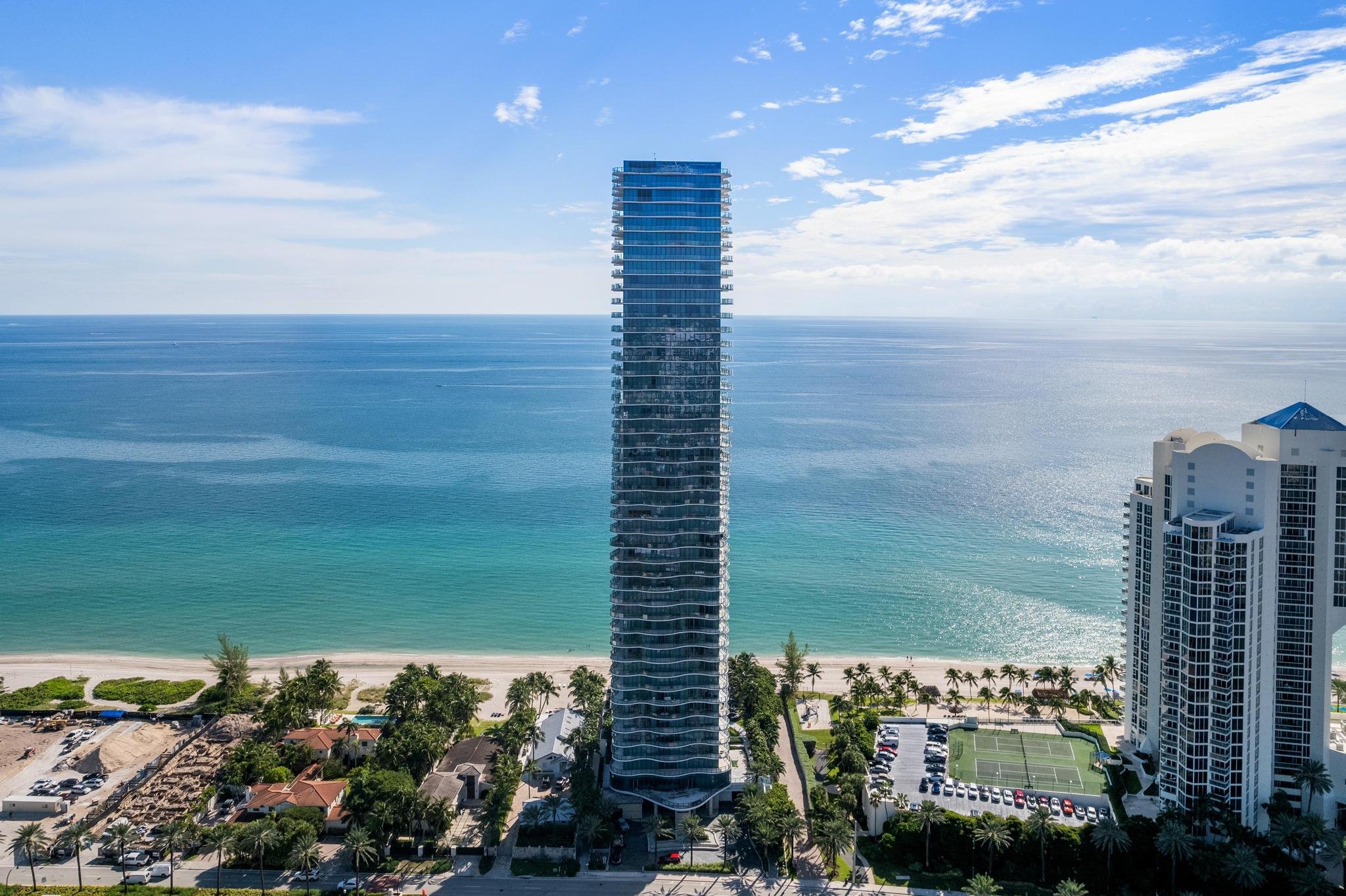 Tall beachfront skyscraper with ocean view, flanked by luxury apartments, palm trees, tennis court, and sandy beach under a blue sky.