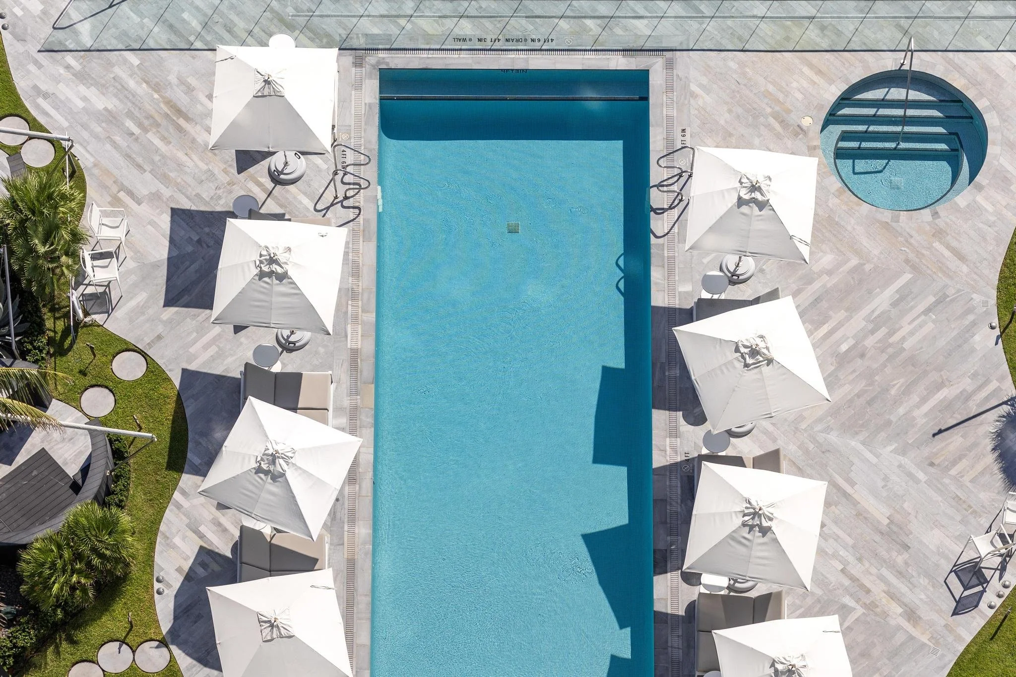 Aerial view of a rectangular swimming pool with surrounding white umbrellas and lounge chairs. Beside the pool, there is a small circular hot tub. The area is tiled and landscaped with grass and trees.