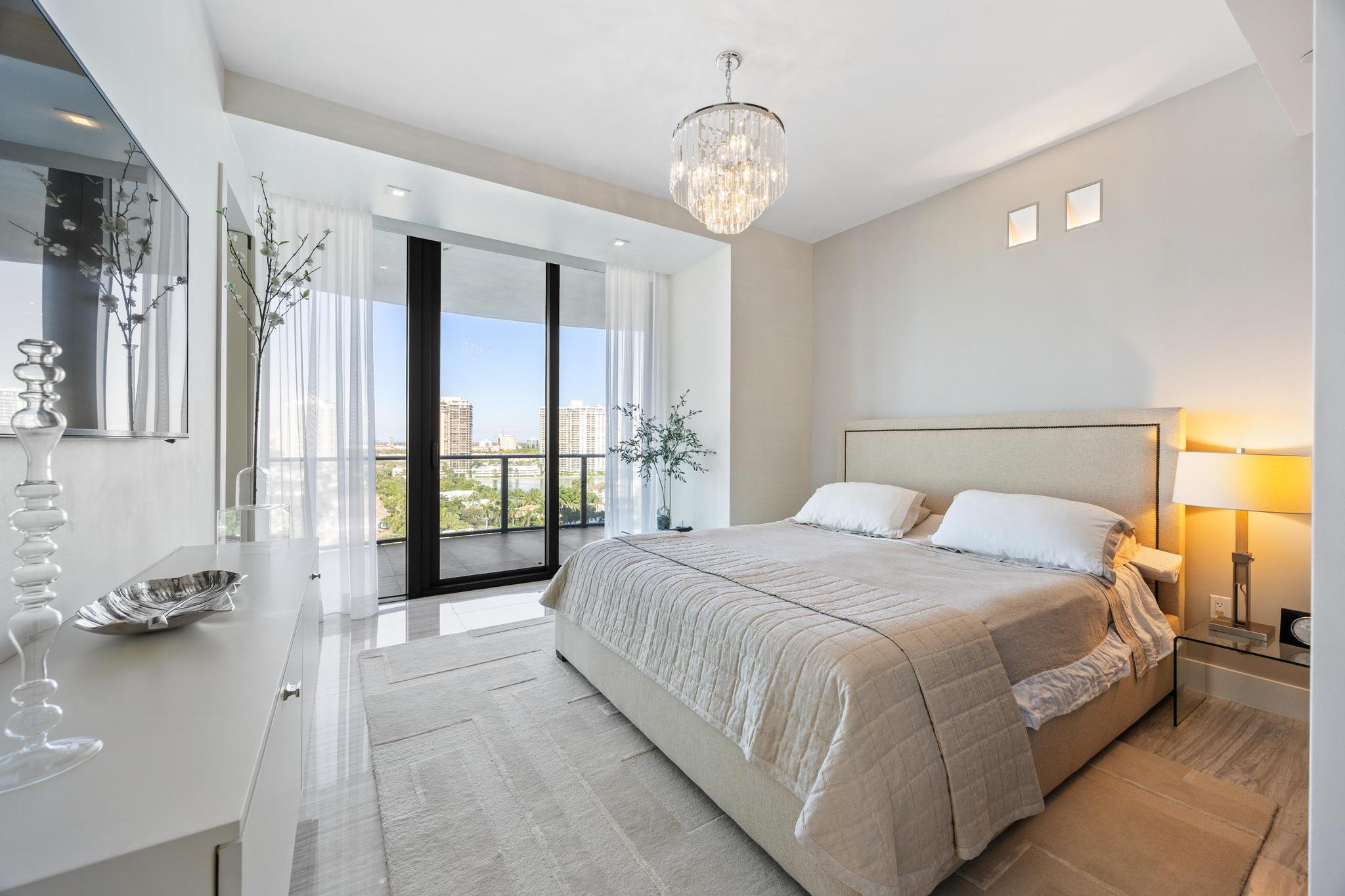 Modern bedroom with large bed, neutral decor, chandelier, balcony view, and minimalistic furniture.