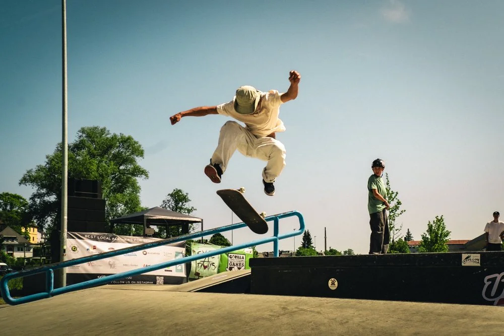 Skateboarder springt über eine Geländer im Skatepark, während andere im Hintergrund zuschauen.
