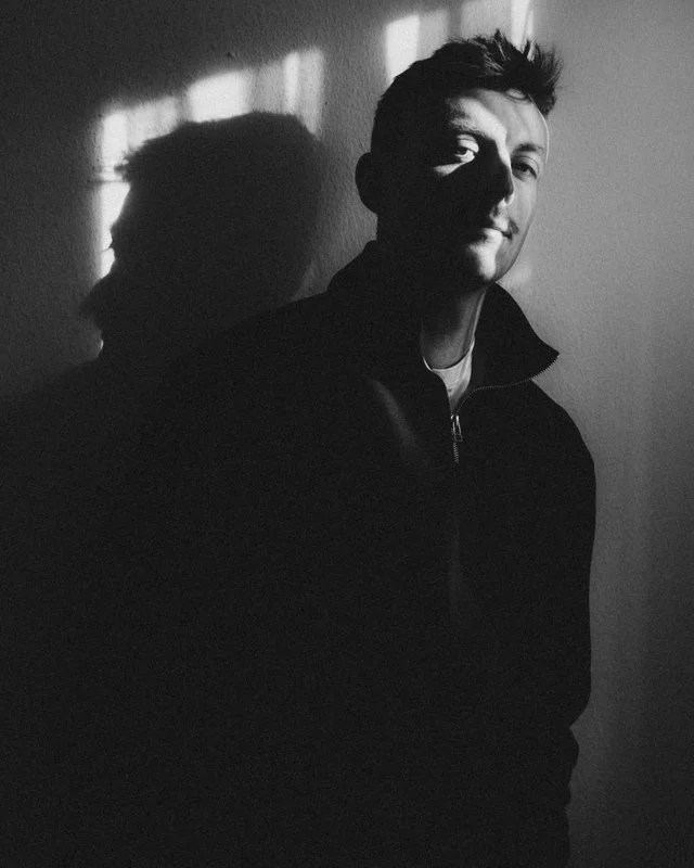 Ein junger Mann steht vor einer Wand, sein Gesicht ist teilweise im Schatten, und er blickt lächelnd in die Kamera. Das Licht fällt auf seine Schulter und sein Gesicht, während sein Schatten an der Wand hinter ihm sichtbar ist.