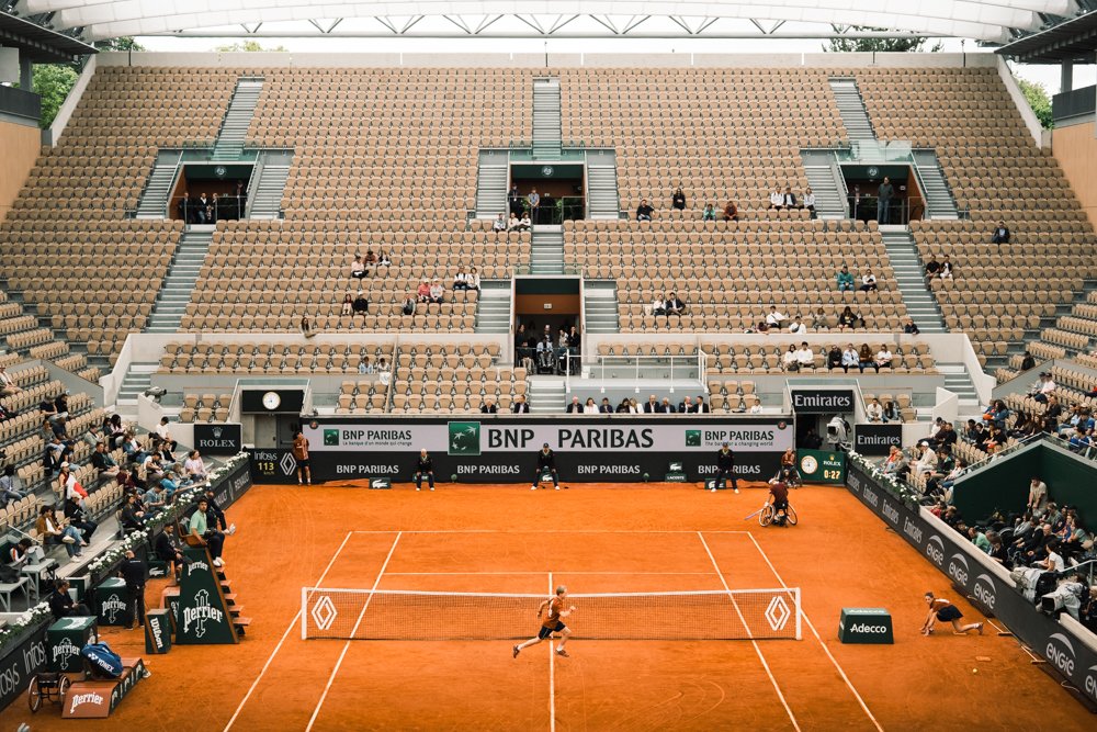 Ein Tennisplatz im Indoor-Stadion mit Spielern, Zuschauern und Werbetafeln auf den Rändern.