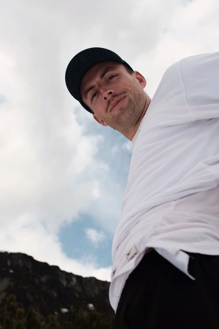 Ein Mann trägt eine schwarze Kappe und ein weißes T-Shirt, steht vor einem bewölkten Himmel und einer Berglandschaft im Hintergrund.