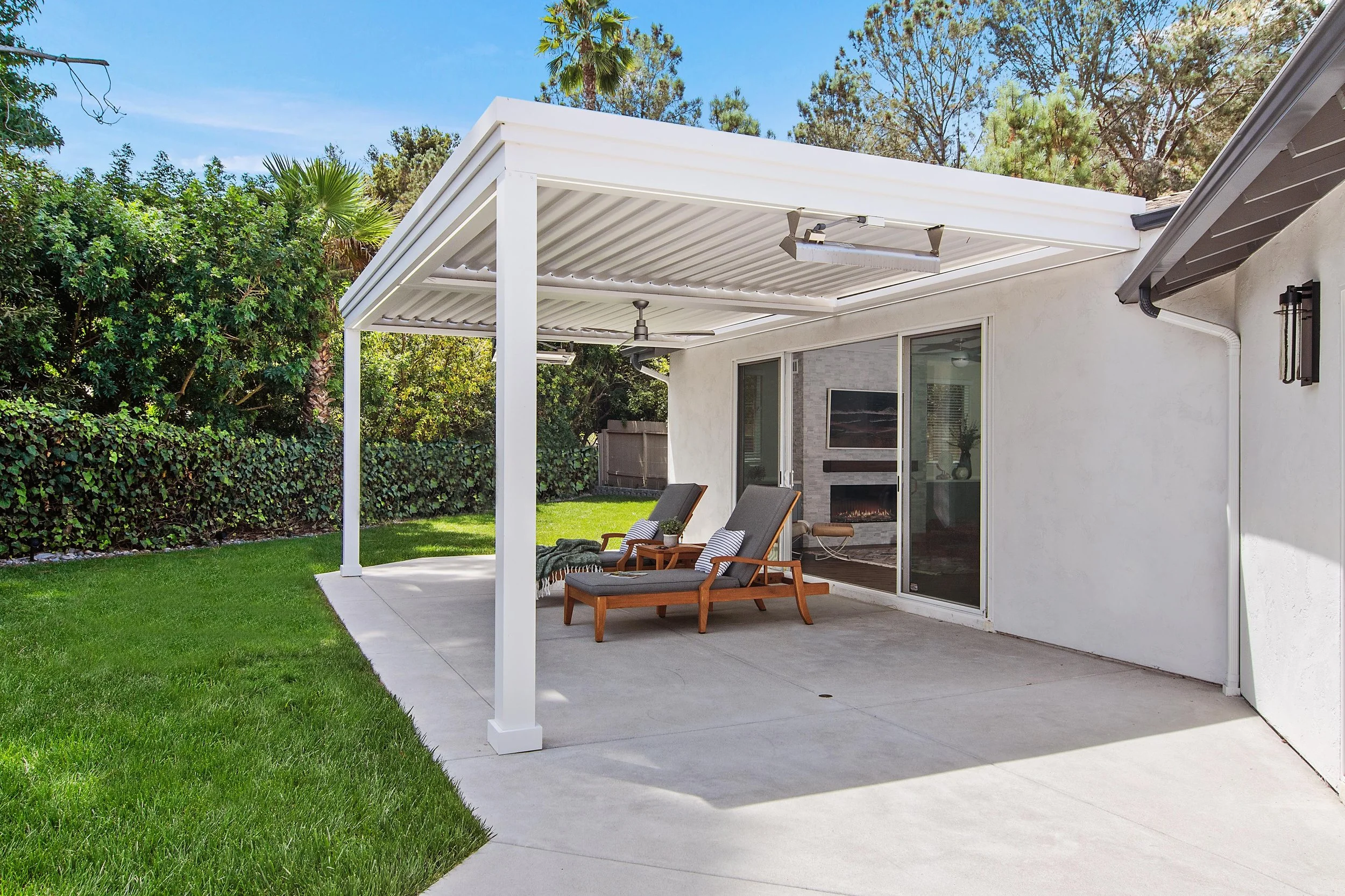 Backyard patio with white pergola, two lounge chairs, a grassy yard, and surrounding trees.
