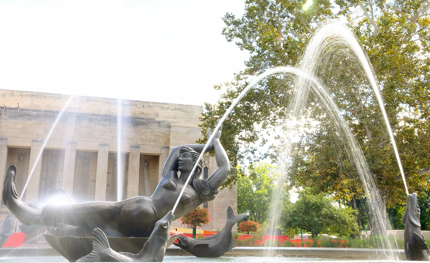Fountain with a statue of a mermaid-like woman holding a large fish, surrounded by other fish sculptures, with water arcing from the fish's mouths, set in a park with trees and a building in the background.