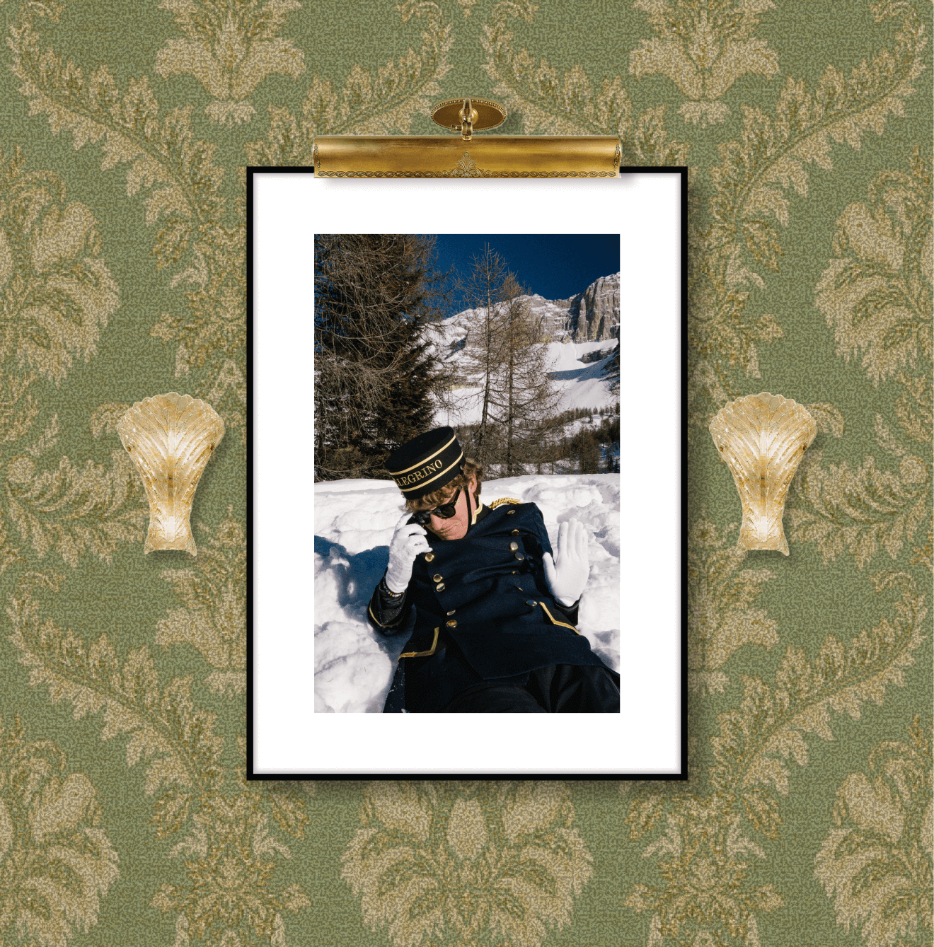 A framed photograph of a person in a dark blue military-style coat and hat, lying in snow with mountains and leafless trees in the background, is hung on a green, gold, and beige floral-patterned wallpaper.