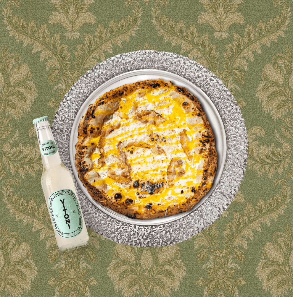 A pizza with a golden and slightly charred crust, topped with cheese and possibly chicken, on an ornate silver platter. A small bottle of Vitoni lemon-flavored aperitif is placed beside the plate, all set on a green patterned tablecloth.