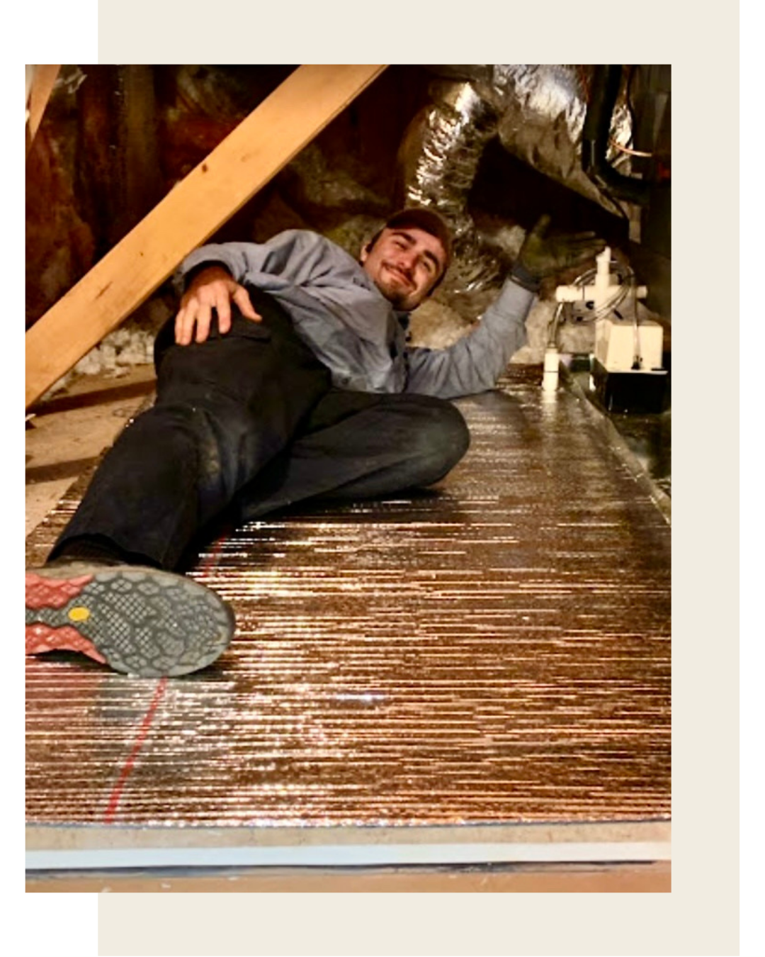 Man lying on his side in an attic or crawl space, smiling, with insulation and ductwork behind him, working near HVAC equipment.