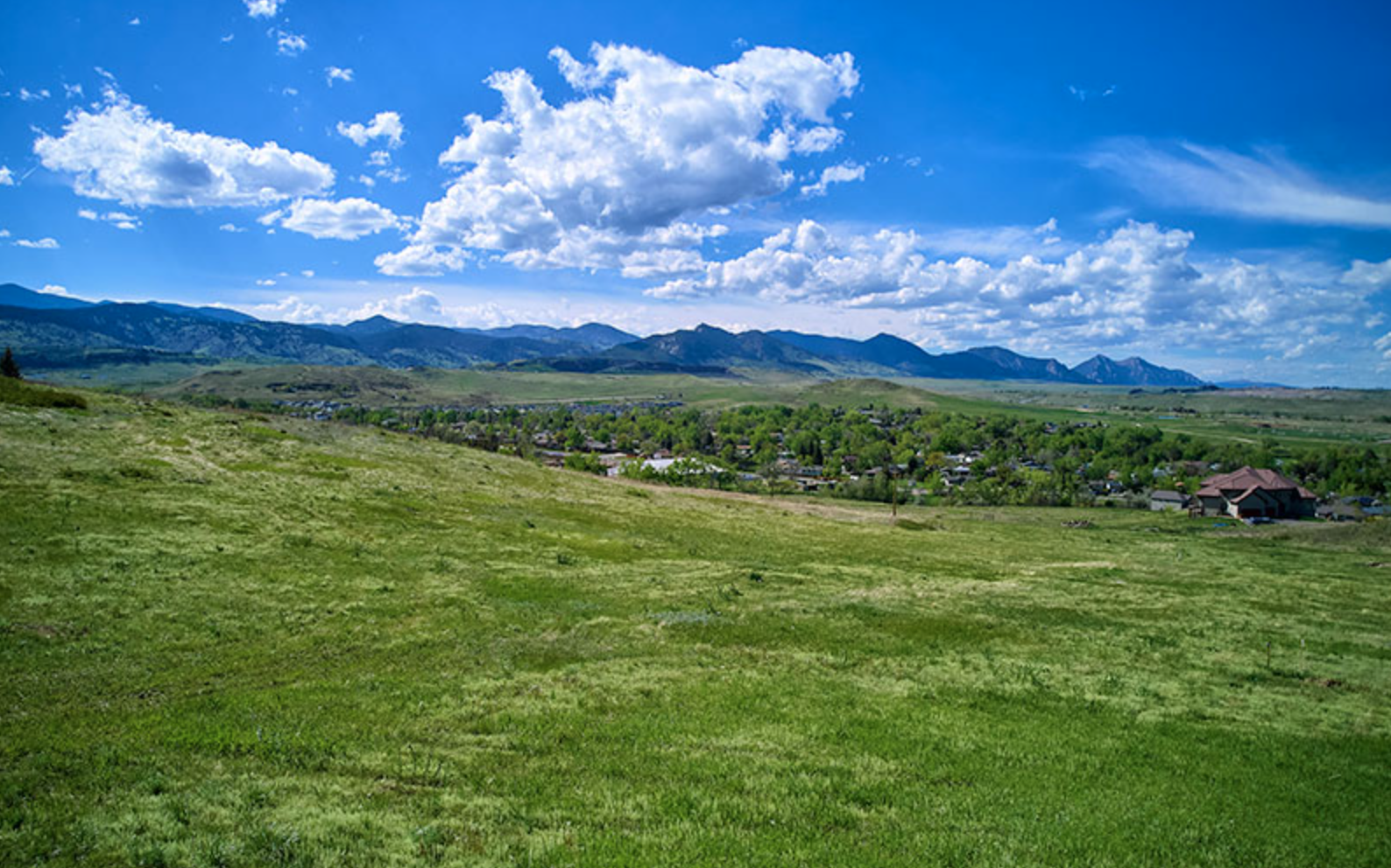 A scenic landscape featuring green grassy fields, a small town, and distant mountains under a bright blue sky with fluffy white clouds.