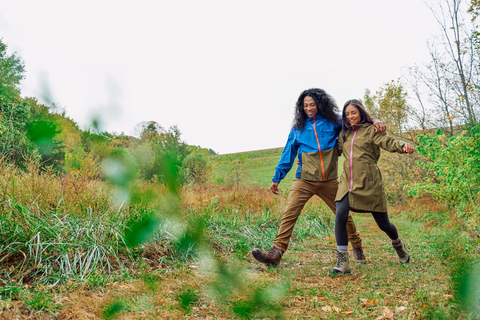 Couple Strides Grassy Fields in Cotopaxi Rainwear.