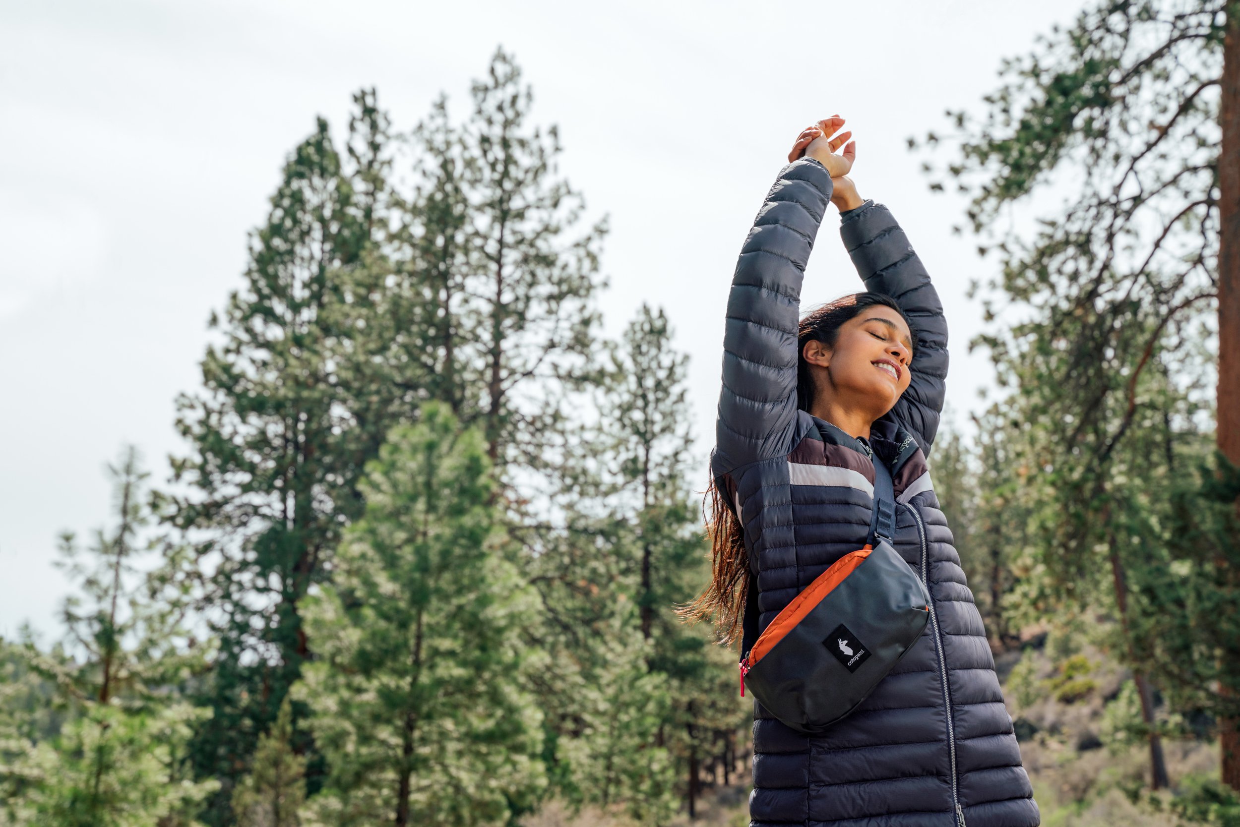 Women stretches among pines with Cotopaxi Kapai Hip Pack and Cotopaxi Fuego Down Parka.