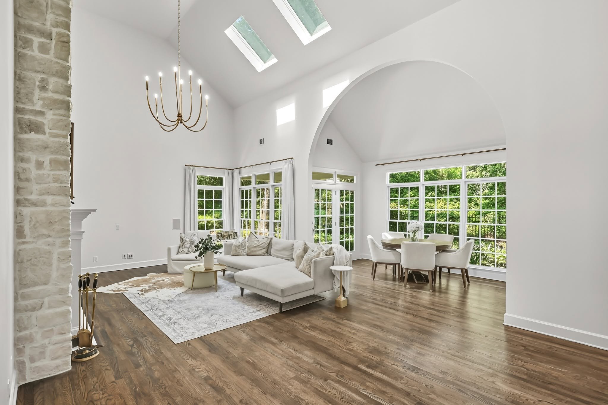 Bright living room with white walls, large windows, wooden floors, a white sectional sofa, a round coffee table, dining table with chairs, a chandelier, and skylights in the ceiling.