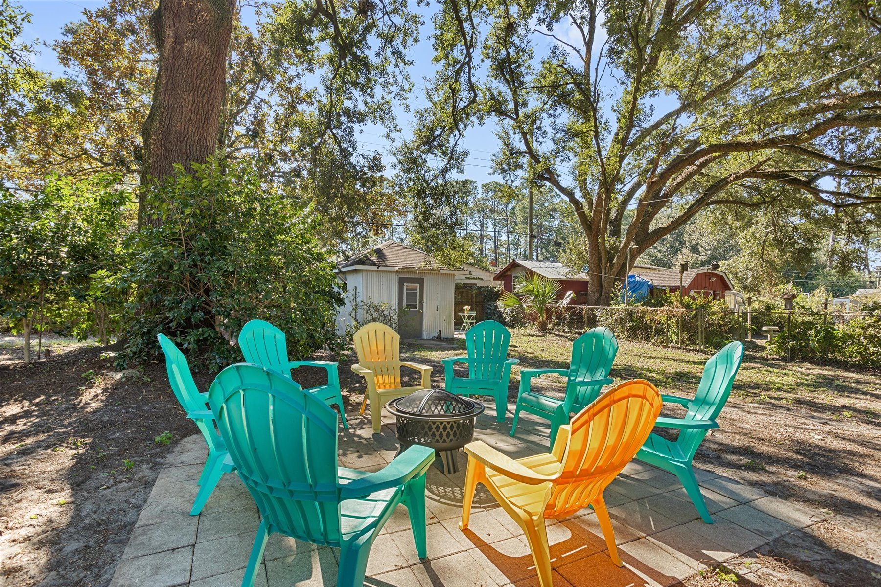 Colorful plastic chairs arranged in a circle around a fire pit in a backyard with a large tree, bushes, and small sheds in the background.