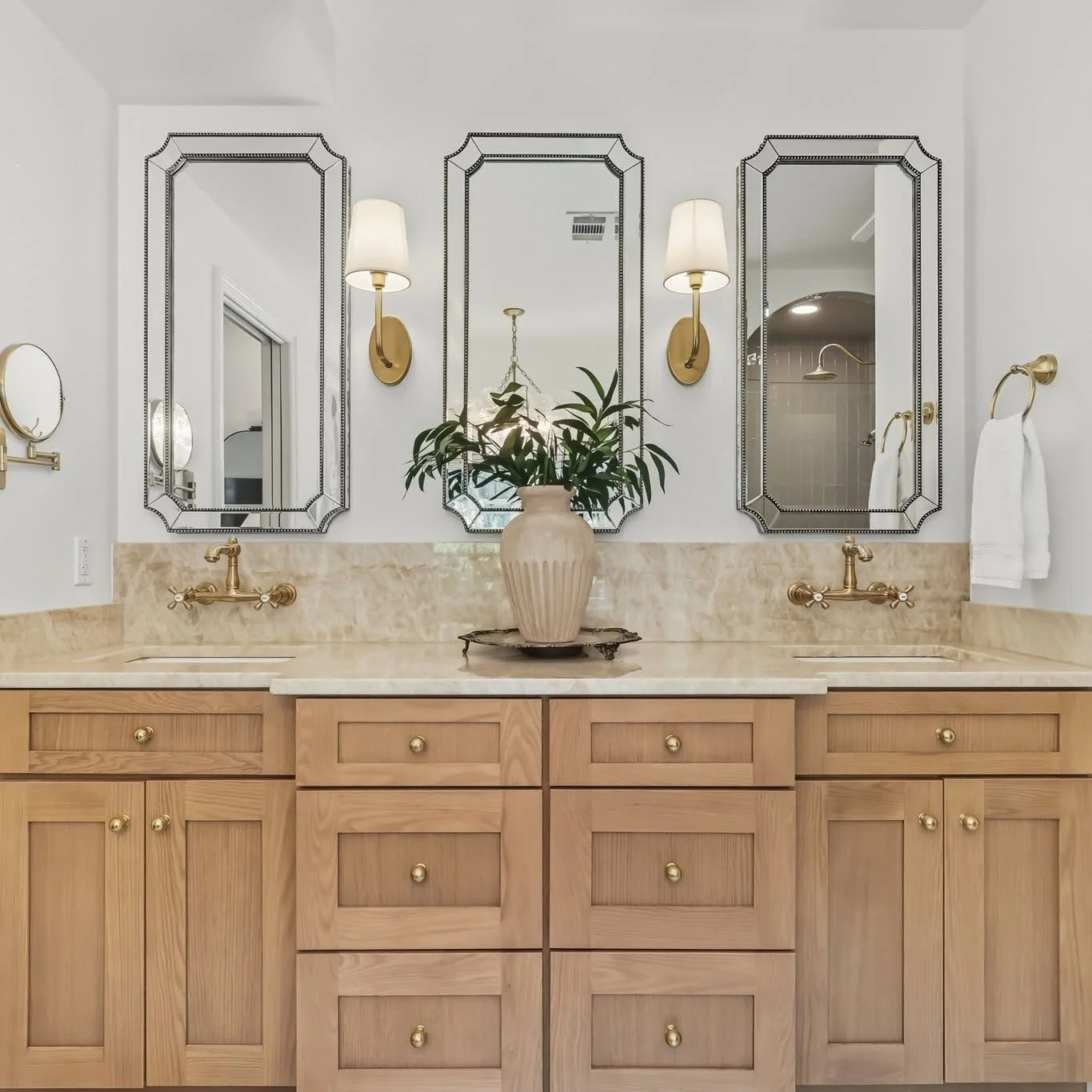 Bathroom with dual sinks, wooden cabinetry, three large mirrors, gold fixtures, white walls, a potted plant on the counter, and a shower area visible through the reflection in the mirrors.