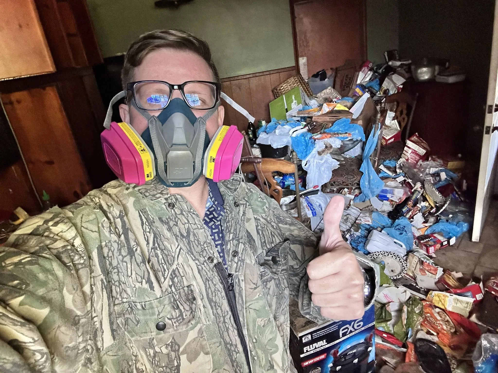 A man wearing camouflage shirt, safety glasses, and pink respirator mask takes a selfie in a cluttered room full of trash and discarded items, giving a thumbs-up gesture.