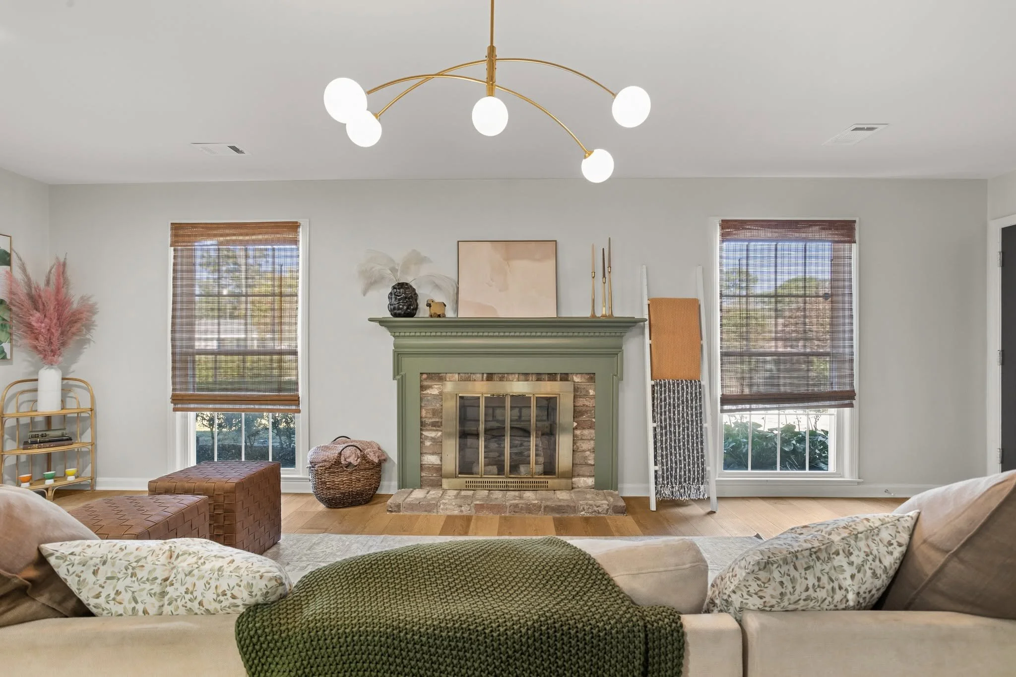 Living room with a fireplace, two large windows with bamboo blinds, and various decorative items including a lamp with white feathers, artwork, and plants.