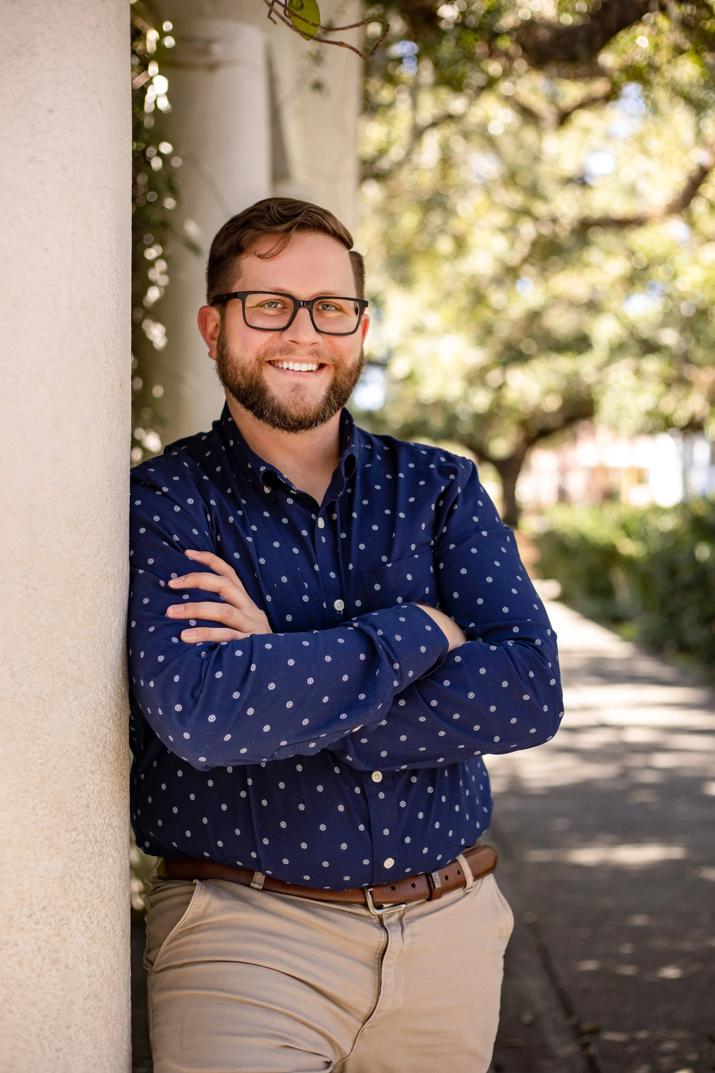 A man with glasses and a beard smiling, leaning against a wall outside on a sunny day, with trees and a sidewalk in the background.