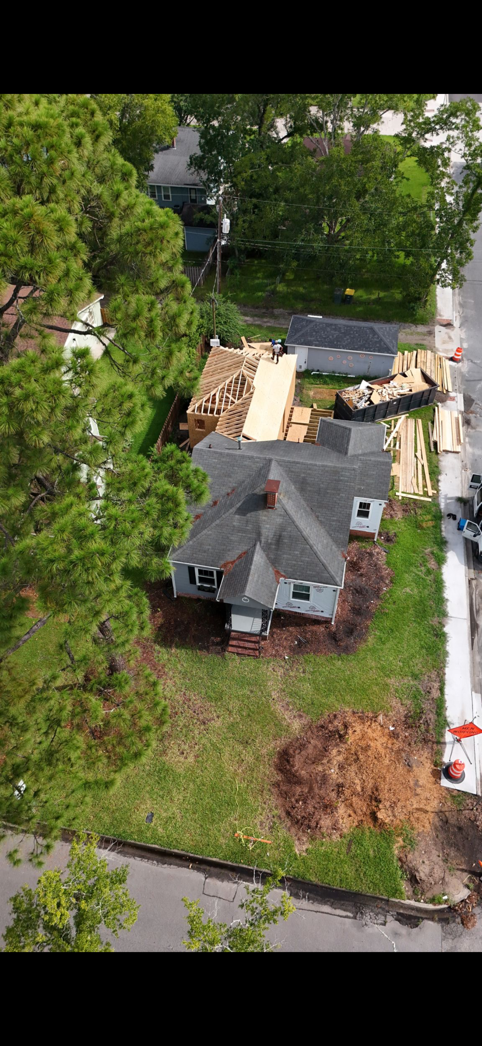 Aerial view of house under construction, with wooden framing, a construction dumpster, and construction workers, surrounded by green trees and a street.