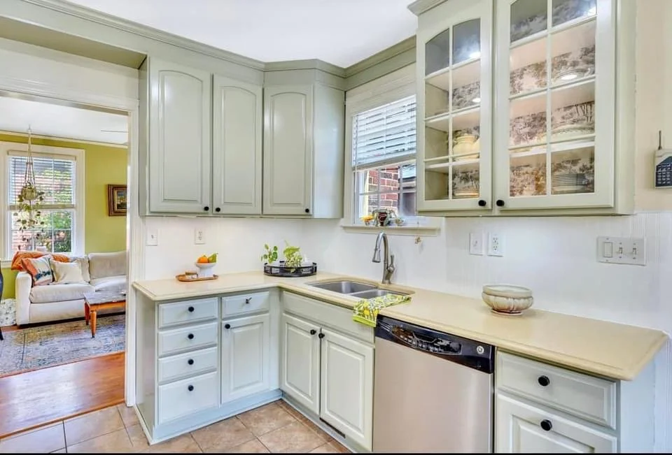 A kitchen with light green cabinets, a window above the sink, and a glass-front cabinet displaying dishes. The counter has a bowl and some small plants, with a dishwasher beneath. Adjacent to the kitchen is a living room with a white couch and yellow