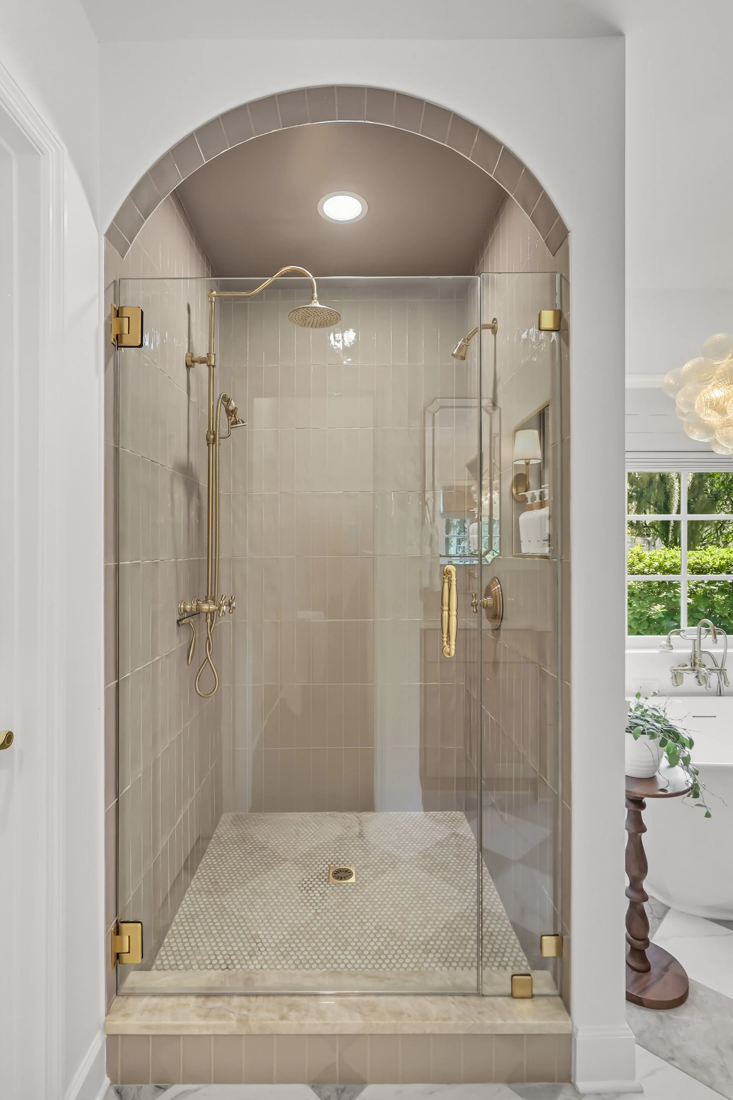 A modern corner shower with glass door, beige tiled walls, gold fixtures, and a small window showing greenery outside.