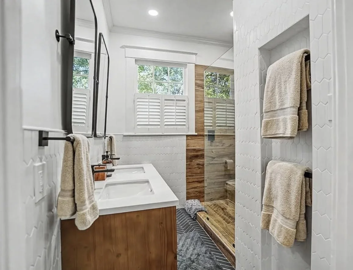 Modern bathroom with dual sinks, large windows with white shutters, towel racks with beige towels, a wood-paneled shower area with a glass door, and a wooden floor in the shower stall.