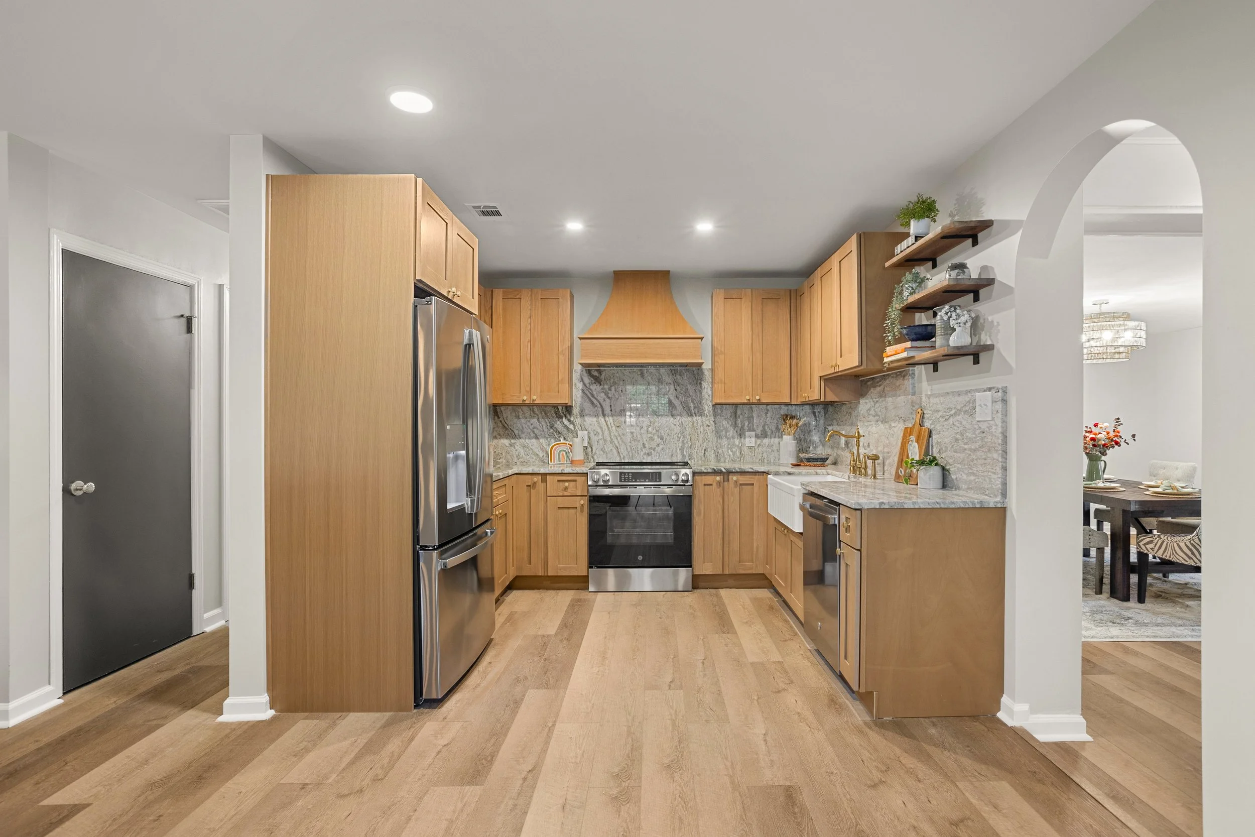 Kitchen with light wood cabinets, stainless steel refrigerator, black oven, and granite countertops. Open living space with dining area in background, decorated with flowers and plants.