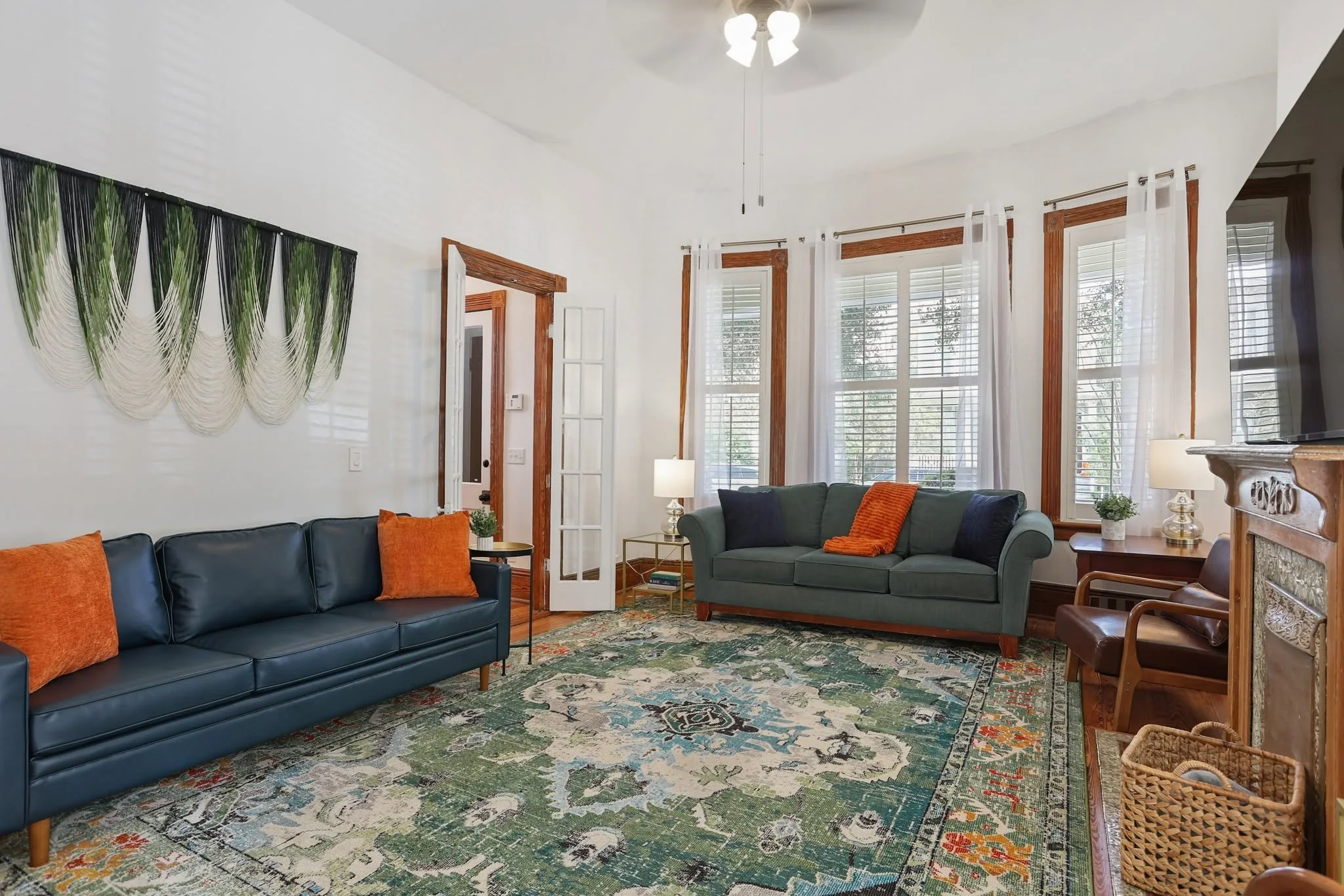 Living room with blue and green sofas, colorful rug, large windows with white curtains, and artwork on the wall.