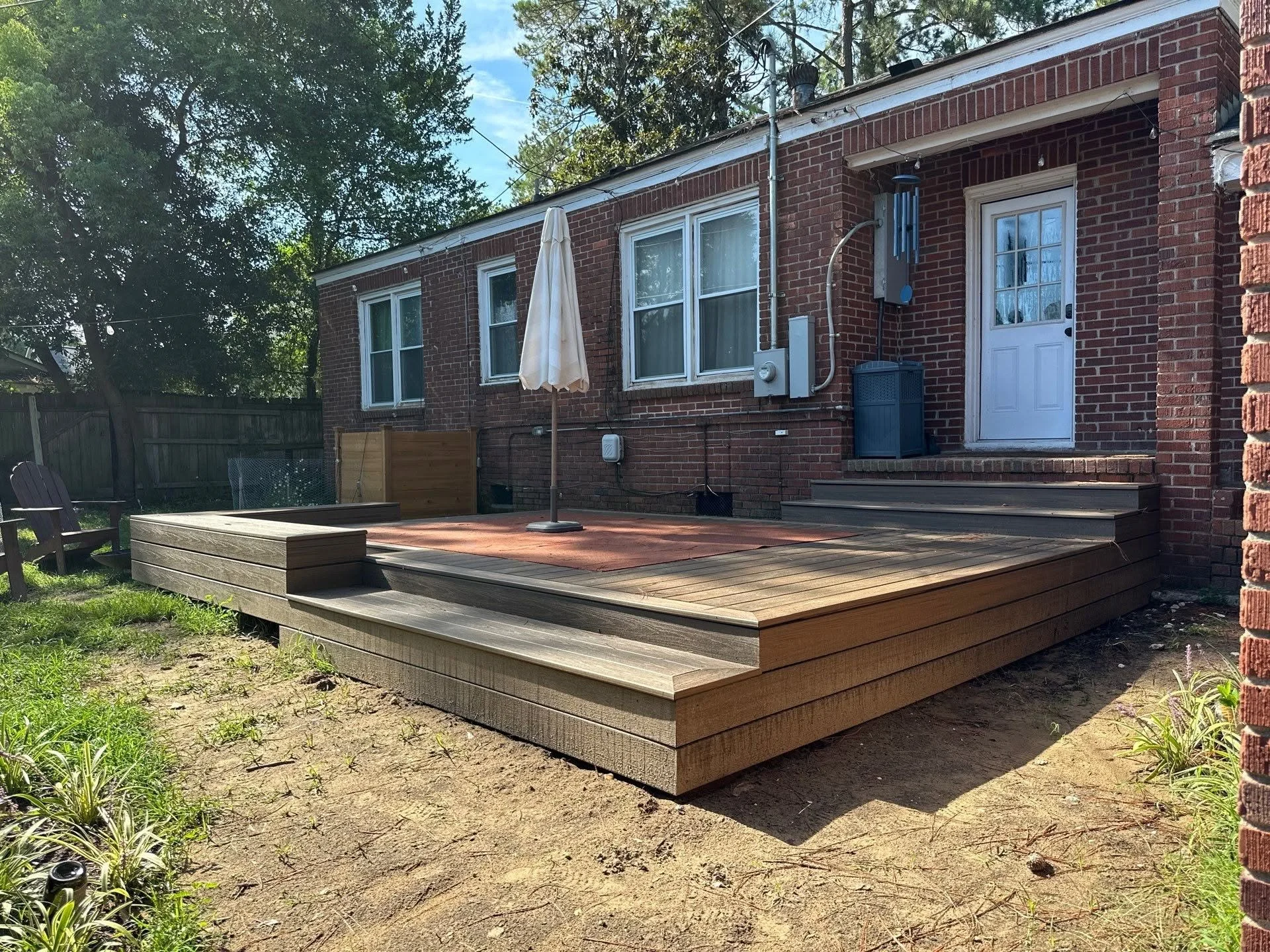 Backyard with a brick house, wooden deck, a closed white door, and a closed patio umbrella. Trees and a wooden fence are in the background.