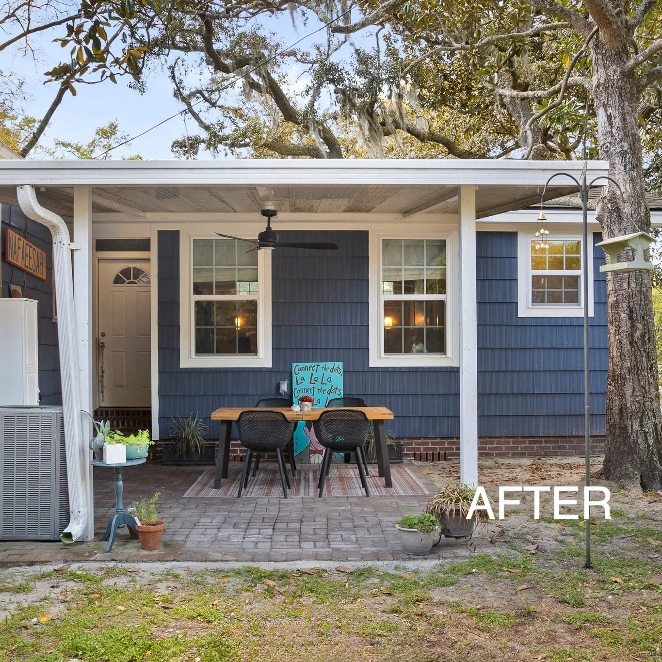 A covered patio with a small table and three chairs in front of a blue house. The table has a blue sign that says ‘Connect the dots’ with some other decorations. There are potted plants on the ground and a hanging birdhouse on the right side of the i