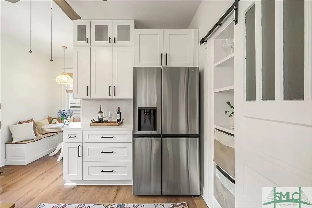 Modern kitchen with white cabinetry, stainless steel refrigerator, and open shelving, with a small seating nook visible on the left.