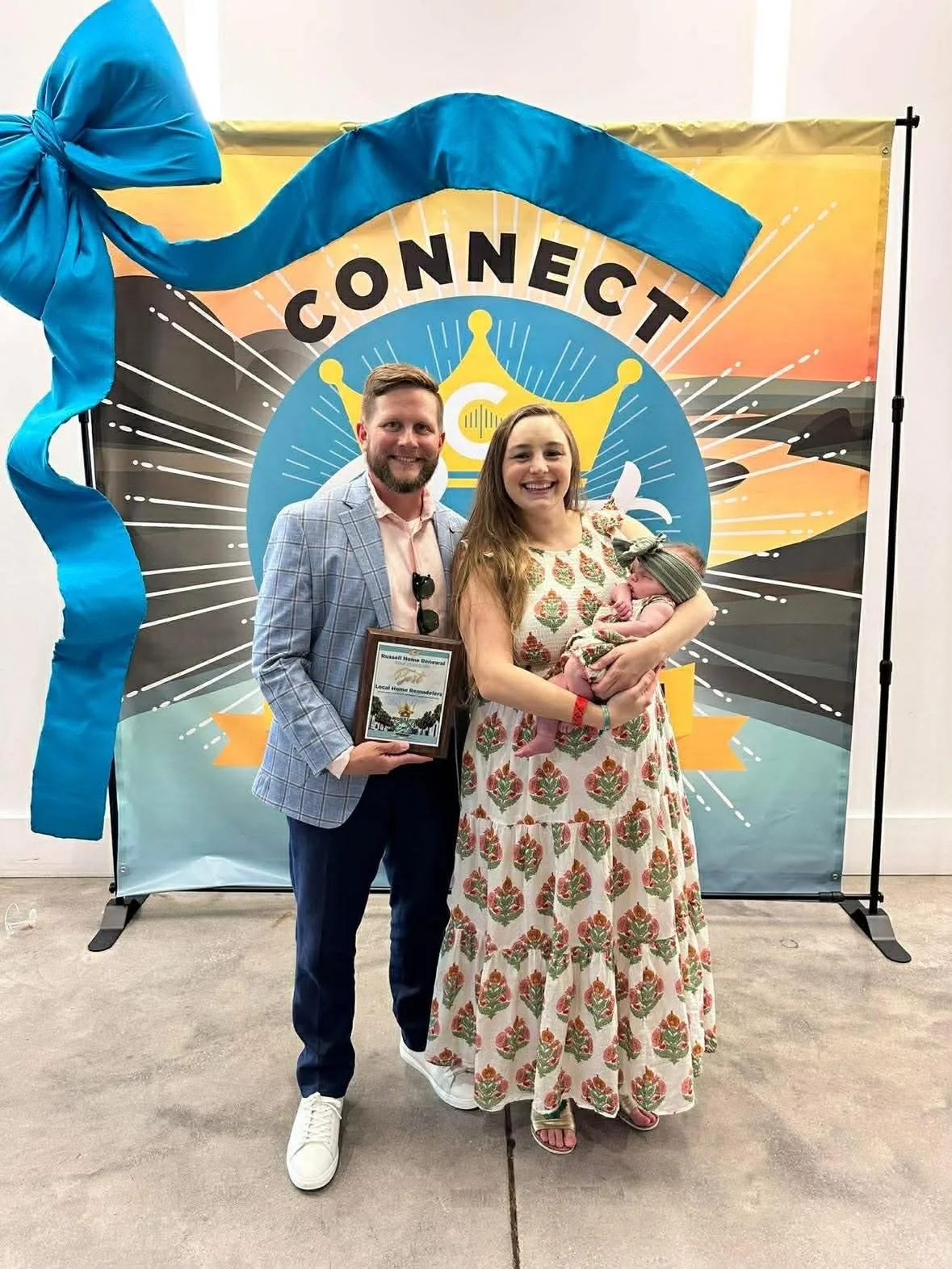 Family celebrating an event at a backdrop with the word 'Connect' and a sunburst design, with a man holding a plaque, a woman holding a baby, and a man with a beard in a plaid blazer.