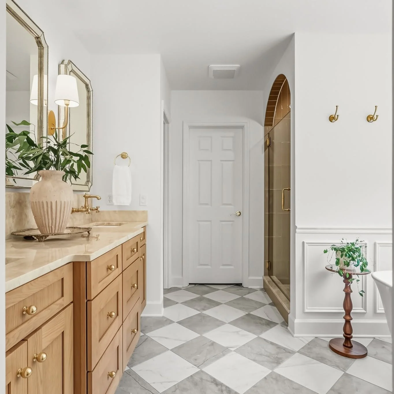 A bathroom with a wooden vanity, marble countertop, large mirror, with a potted plant on it. There is a white door, a glass shower enclosure with gold fixtures, and another plant on a stand near the wall.
