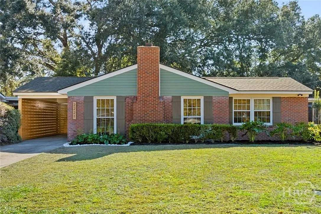 A single-story brick and siding house with a pitched roof, front yard, and a driveway on the left, surrounded by trees in the background.
