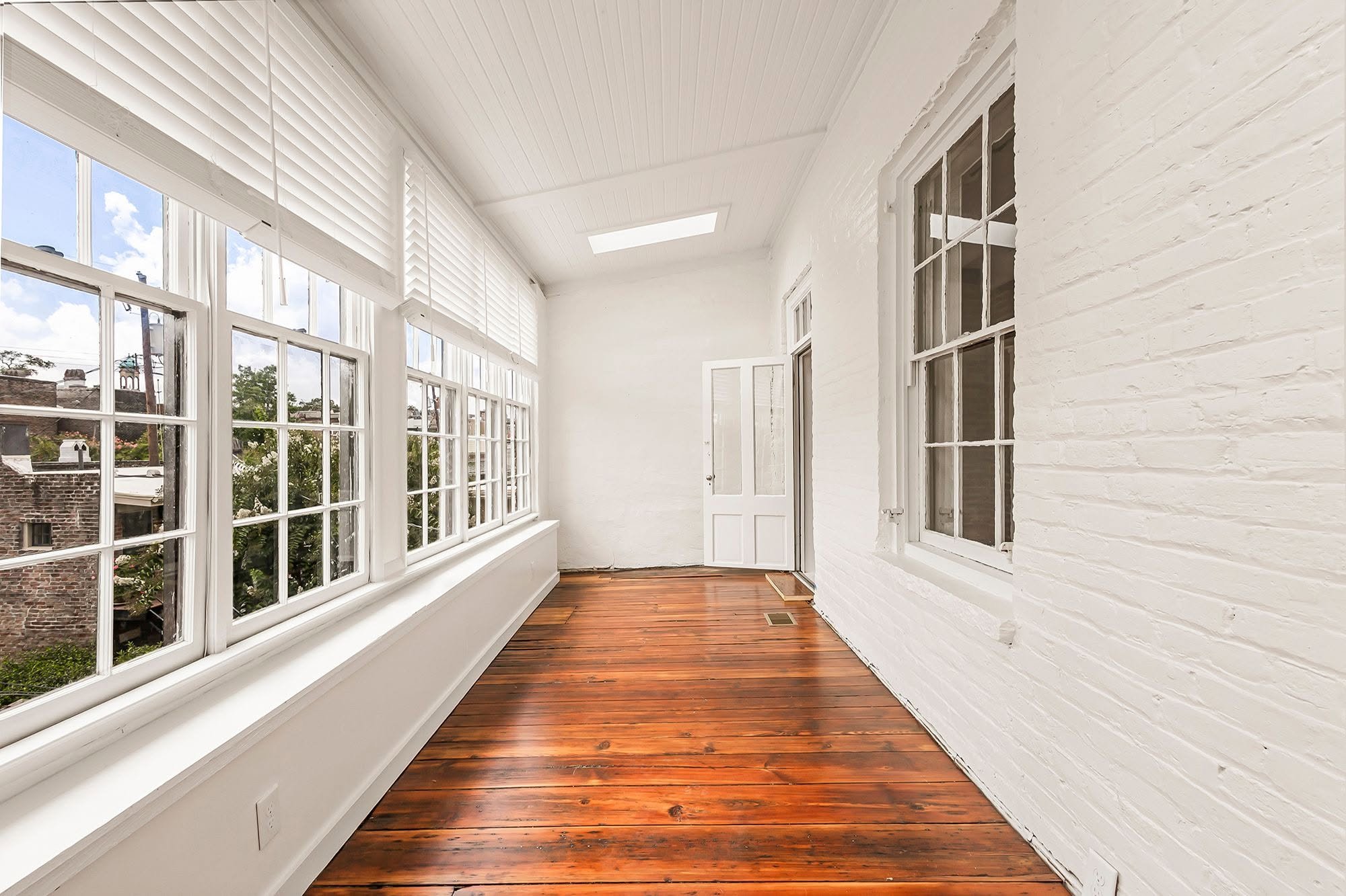 Sunlit enclosed porch with large windows, white painted brick walls, wooden floor, and a skylight in the ceiling.