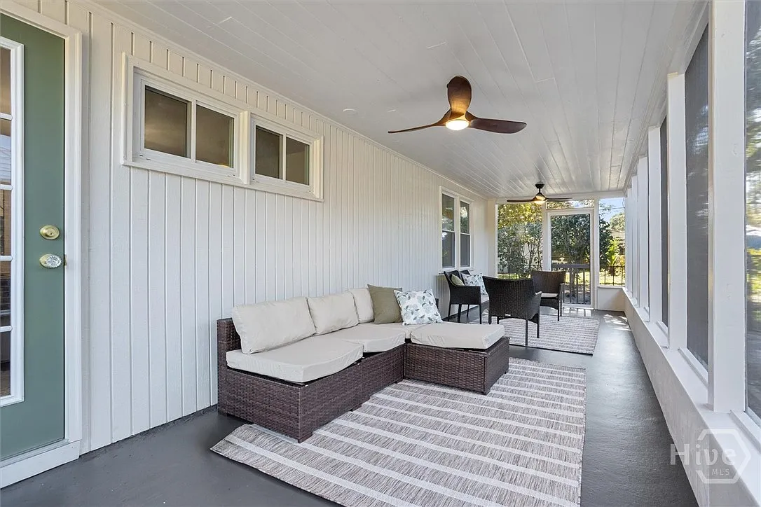 Enclosed porch with white paneled walls, ceiling fans, a wicker sofa with cushions, a rug, and a table with black chairs.